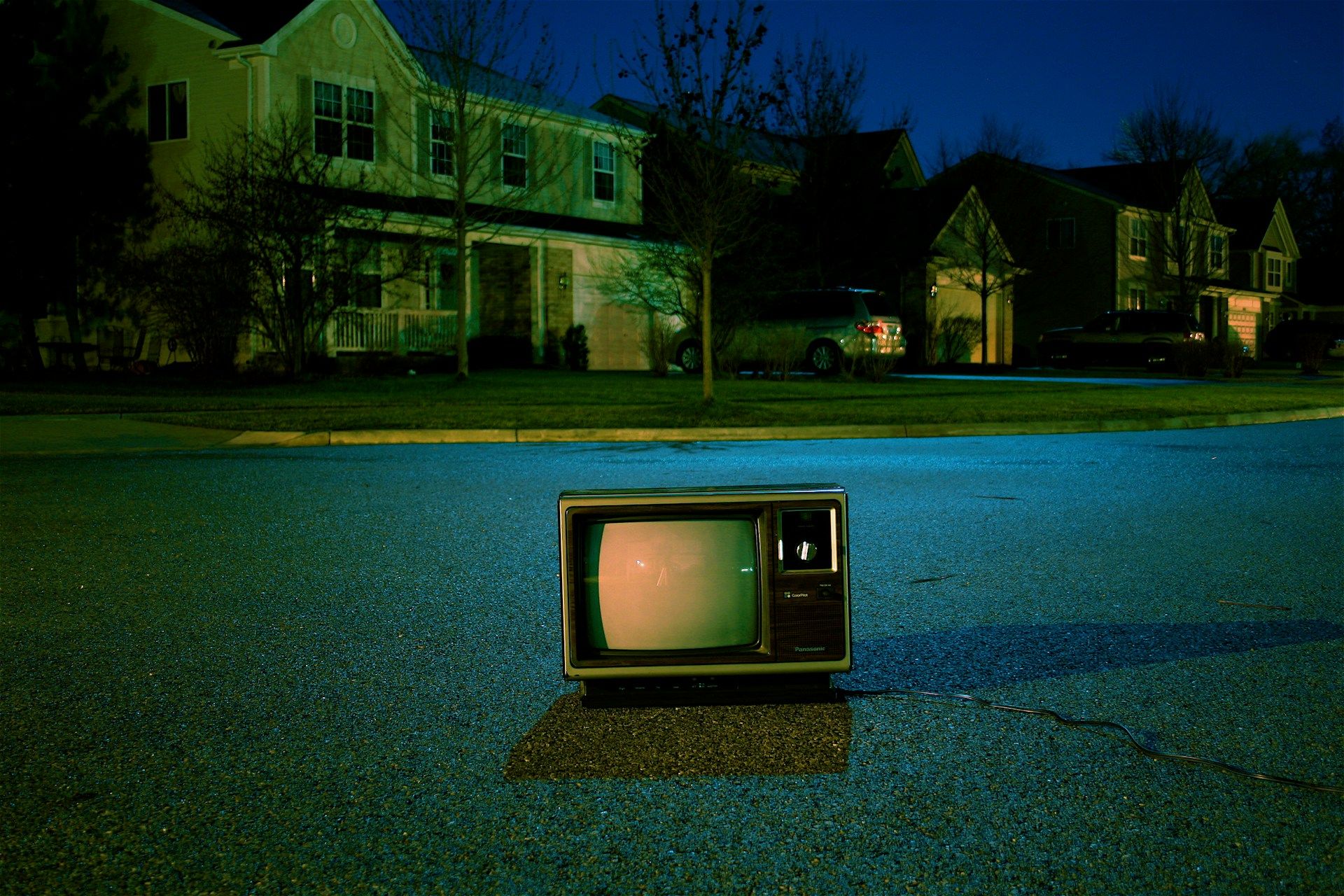 A vintage CRT television in the middle of a suburban road, with houses in the background.