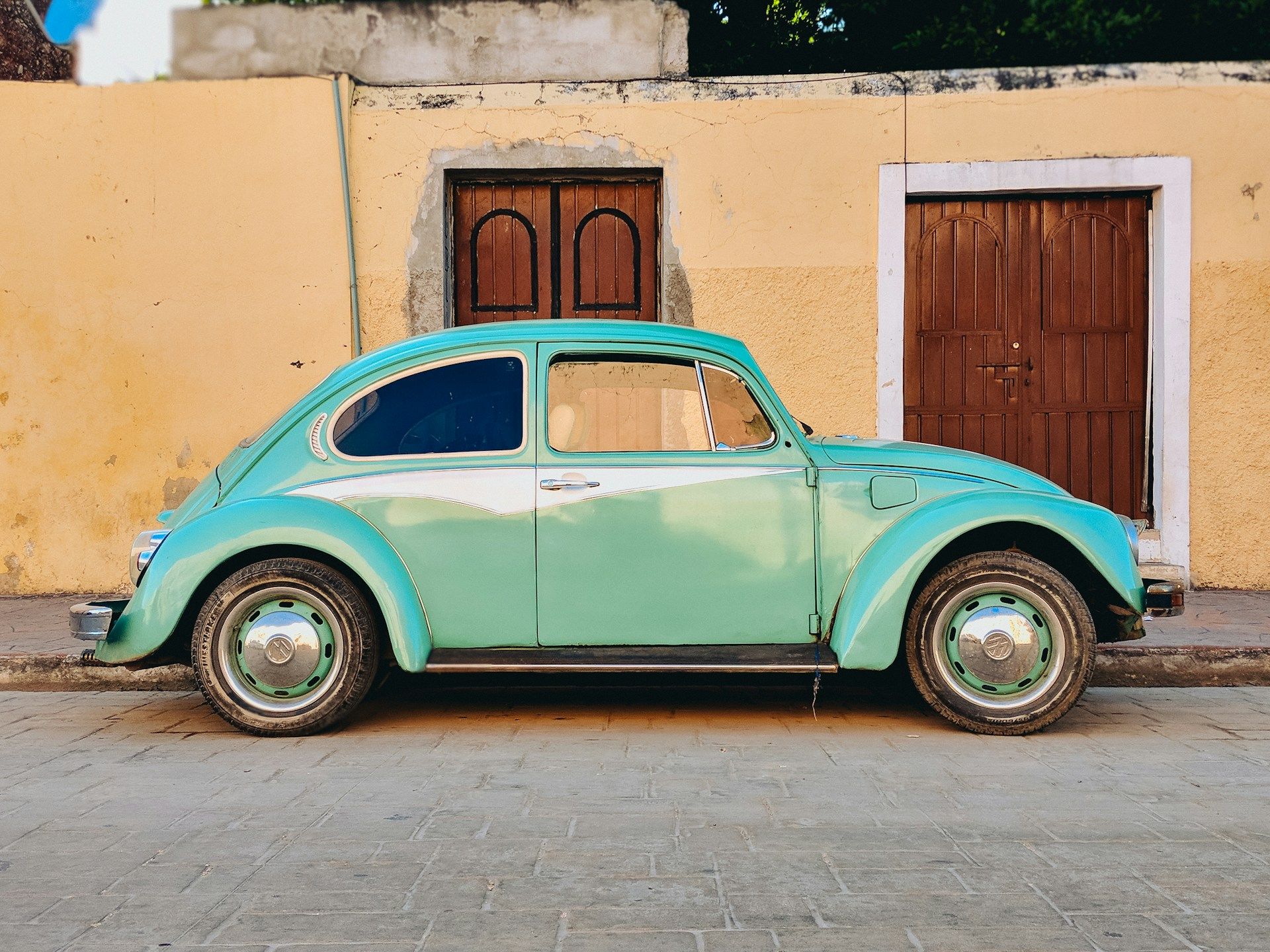 A light blue Volkswagen Beetle, parked in front of a brown house with dark brown doors. 